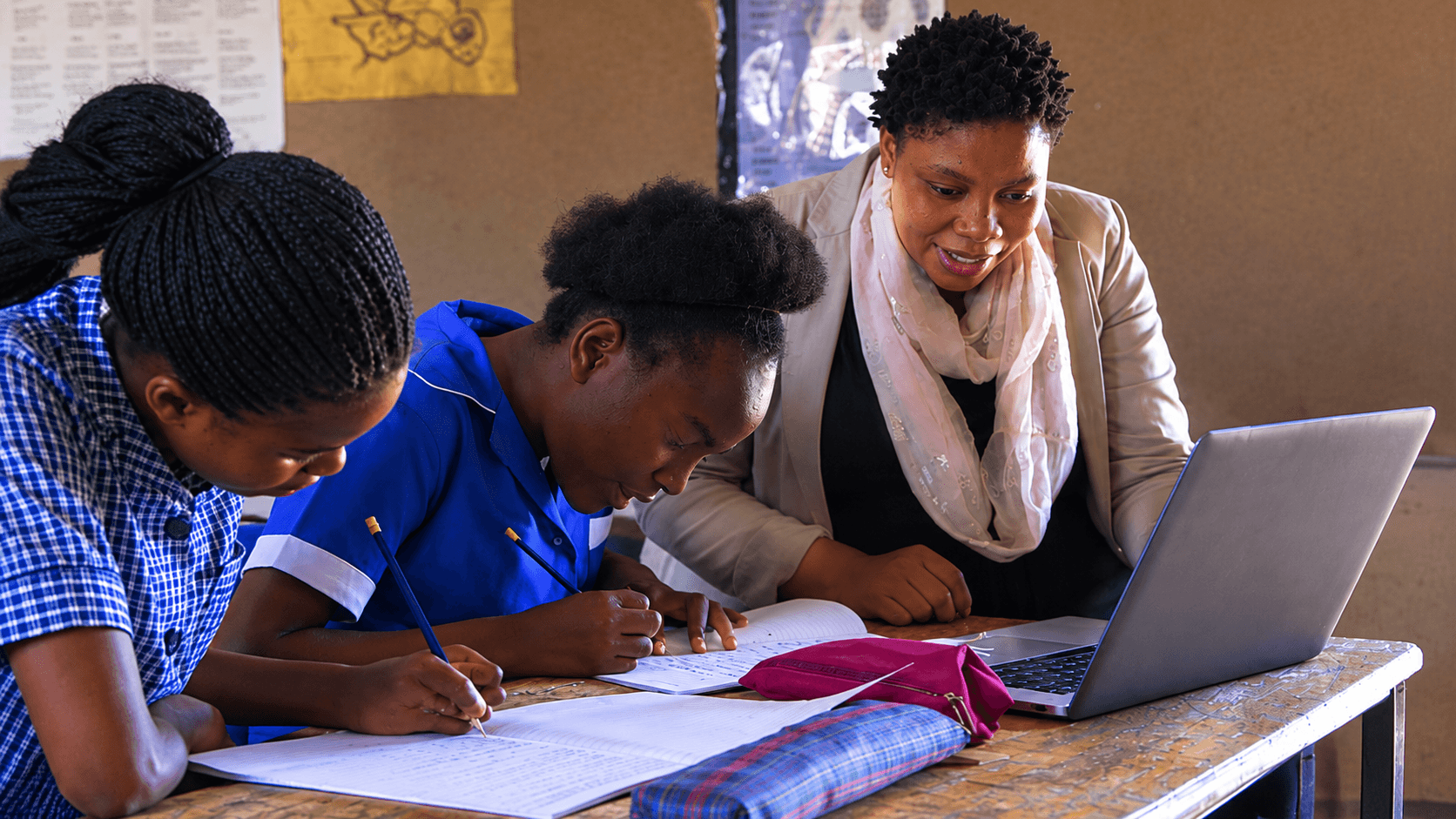 A teacher works on a laptop beside two students in blue school uniforms
